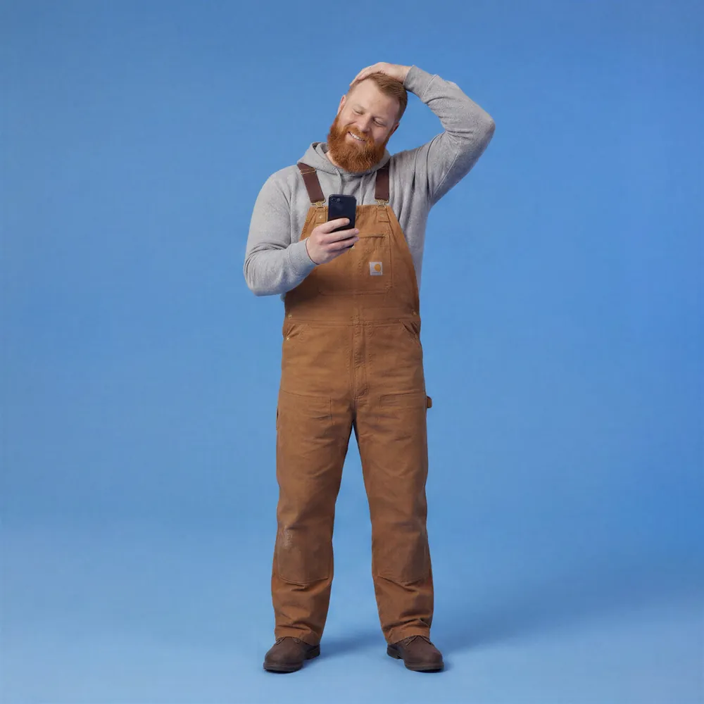 Smiling red-haired man in brown work overalls stretches his neck while looking at a smartphone against a plain blue background.